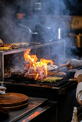 Steaks and meats grilling over bright open flames and smoke on a commercial kitchen grill.