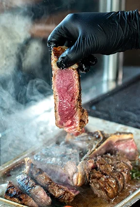Gloved hand placing a thick cut of raw beef steak onto a smoking hot outdoor grill.