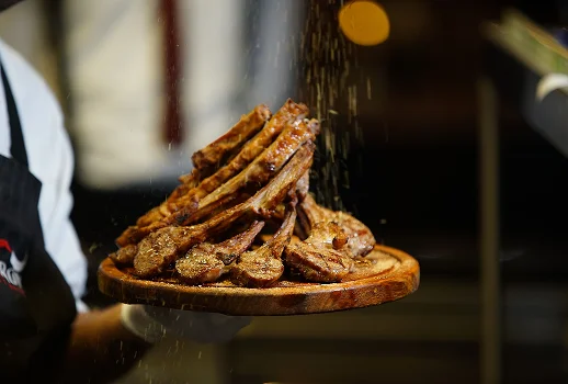 Chef sprinkling coarse salt over a tall stack of cooked lamb chops presented on a wooden board.