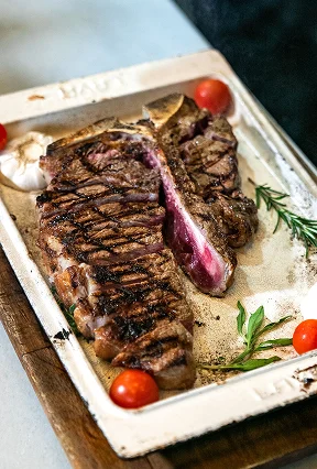 Juicy grilled T-bone steak served on a white platter garnished with cherry tomatoes and rosemary.