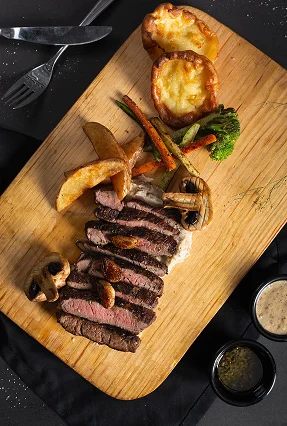 Top-down view of sliced medium-rare beef steak with crispy fries and dipping sauces on a wooden board.