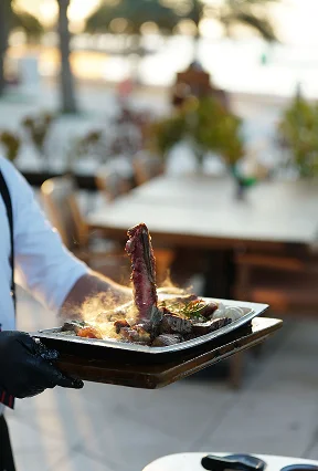 Restaurant waiter carrying a freshly cooked steak platter outdoors in a fine dining setting.